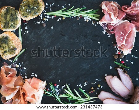 Wine meat appetizer set: prosciutto, serrano and cured lamb meat selection with spices, herbs and baguette slices with pesto on a dark stone background with a copy space in the center