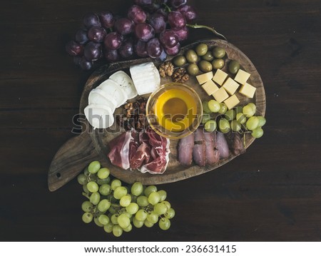 Wine appetizers set: meat and cheese selection, honey, grapes, walnuts and olives on a rustic wooden board over a dark wood background. Top view