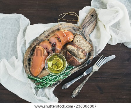 Appetizer set: chicken carpaccio, bread, olives, spices and rosemary on a rustic wooden board and a piece of linen fabric over a dark wood background. Top view