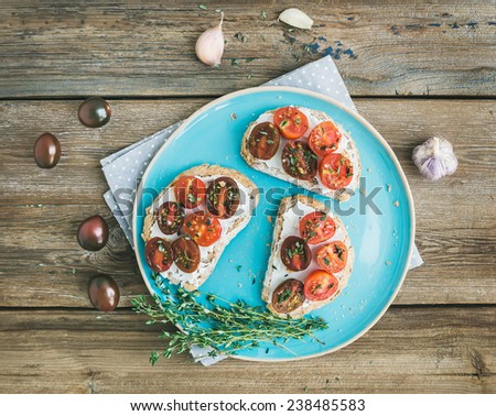 Rustic breakfast set of sandwiches with soft cream-cheese, cherry-tomatoes roasted with garlic and thyme on a rough wood background. Top view