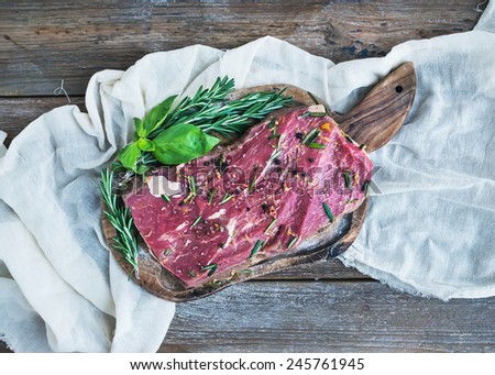 A piece of raw fresh beef (Ribeye steak) marinated in spices and herbs on a rustic wooden board over a kitchen towel and rough wooden desk. Top view