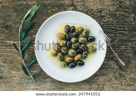 A plate of Mediterranean olives in olive oil with a branch of olive tree and a small silver fork over a rough old wooden desk. Top view