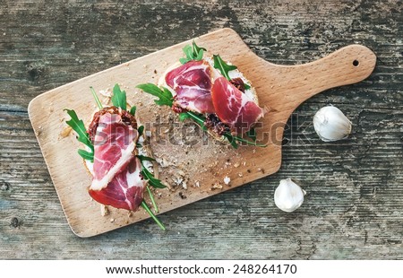 Brusquette set with smoked meat, arugula and dried tomatoes on a rustic wooden board over a rough wood background. Top view