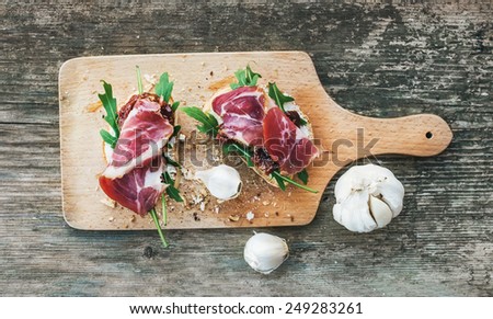Brushetta set with smoked meat, arugula, garlic and dried tomatoes on a rustic wooden board over a rough wood background. Top view
