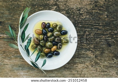 A plate of Mediterranean olives in olive oil with a branch of olive tree over a rough old wooden desk with a copy space. Top view