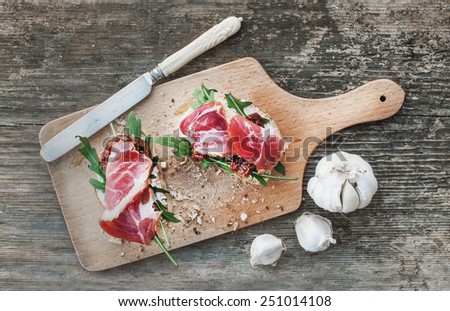 Brushetta set with smoked meat, arugula, garlic and dried tomatoes on a rustic wooden board over a rough wood background. Top view