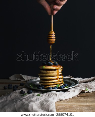 Pancake tower with fresh blueberry and mint on a rustic metal plate, hand is keeping wooden stick pouring honey flow