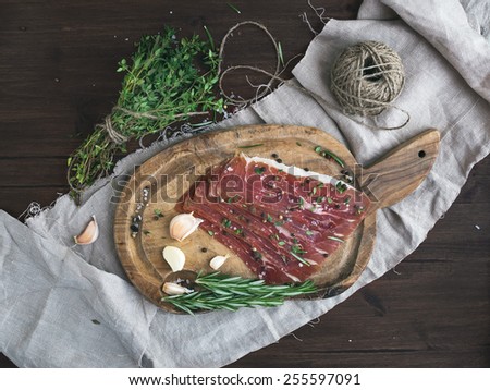 Cured pork meat or prosciutto on a rustic wooden board with garlic, spices and thyme over a piece of linen fabric and dark wood background. Top view