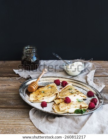 Thin pancakes or crepes with fresh raspberry, cream, mint, on a rustic wooden desk with pomegranate and honey at the backdrop. Black background