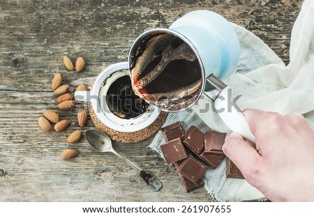 Hand pouring freshly brewed coffee from cezve (coffee pot) to a cup. Roasted almonds, a spoon, a towel, chocolate bar pieces and rough wooden desk at the background. Top view