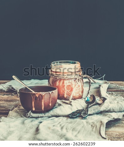 Salted caramel sauce in a rustic glass jar and brown ceramic cup on a wooden desk. Black background, pastel edition