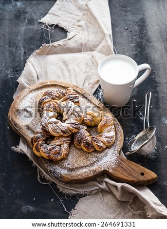 Cinnamon buns with sugar powder on a rustic wooden board, mug of milk, on dark grunge backdrop