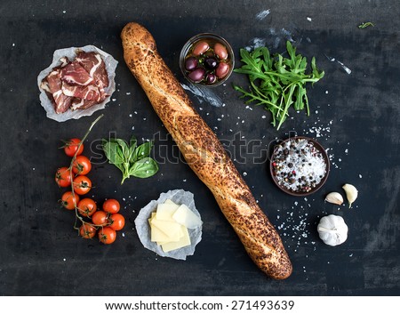 Ingredients for sandwich with smoked meat, baguette, basil, arugula, olives, cherry-tomatoes, parmesan cheese, garlic and spices over black grunge background. Top view.