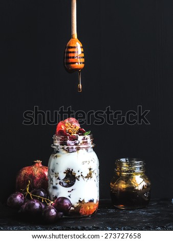 Yogurt and oat granola with grapes, pomegranate and grapefruit in a tall glass jar on black backdrop. Hand pours honey over the jar with a wooden stick