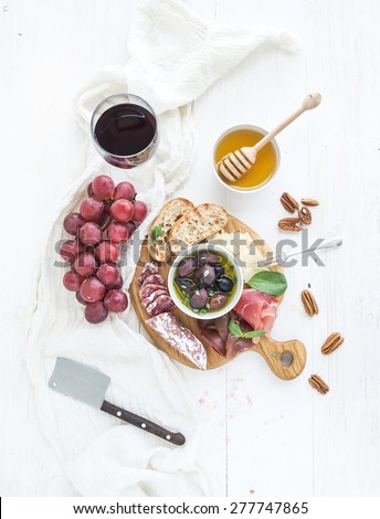 Wine appetizer set. Glass of red wine, grapes, honey, parmesan cheese, meat variety, bread slices, pecan nuts, honey, olives and basil on rustic wooden board over white wood backdrop. Top view