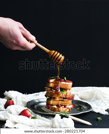 French toasts tower with strawberry, cream cheese and mint, hand is pouring honey over top of it. Dark background