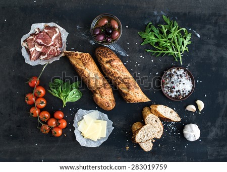 Ingredients for sandwich with smoked meat, baguette, basil, arugula, olives, cherry-tomatoes, parmesan cheese, garlic and spices over black grunge background. Top view