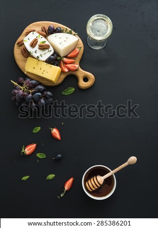 Cheese appetizer selection or whine snack set. Variety of cheese, grapes, pecan nuts, strawberry and honey on round wooden board over black backdrop, top view, copy space