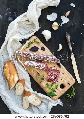 Meat gourmet snack. Salami, garlic, baguette and herbs on rustic wooden board over dark grunge backdrop, top view