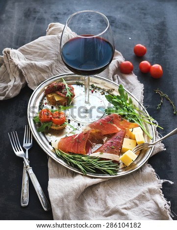 Wine appetizer set. Glass of red wine, vintage dinnerware, brushetta with cherry, dried tomatoes, arugula, parmesan, smoked meat on silver tray over rustic grunge surface. Closeup