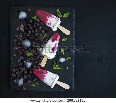 Black-currant and cream ice-creams or popsicles with frozen black-currant, ice cubes and mint on black slate tray over dark grunge backdrop, top view, copy space