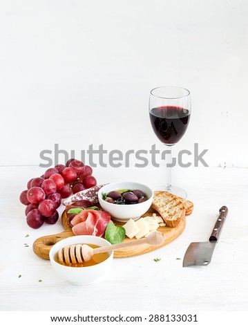 Wine appetizer set. Glass of red wine, grapes, honey, parmesan cheese, meat variety, bread slices, pecan nuts, honey, olives and basil on rustic wooden board over white wood backdrop