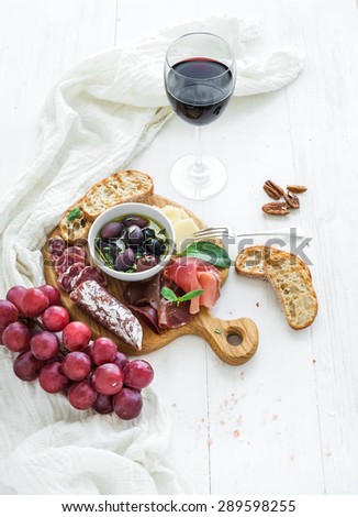 Wine appetizer set. Glass of red wine, grapes, honey, parmesan cheese, meat variety, bread slices, pecan nuts, honey, olives and basil on rustic wooden board over white wood backdrop, top view