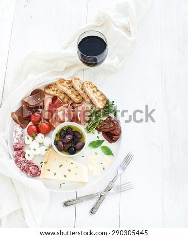 Wine appetizer set. Cherry-tomatoes, parmesan cheese, meat variety, bread slices, dried tomatoes, olives and basil on round ceramic plate over white wood backdrop, top view, copy space