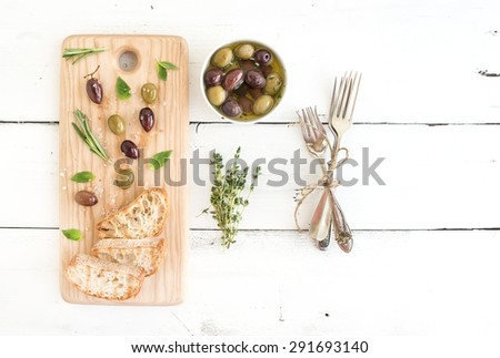 Mediterranean olives with herbs and ciabatta slices on rustic wooden board  over white wooden background, top view, copy space