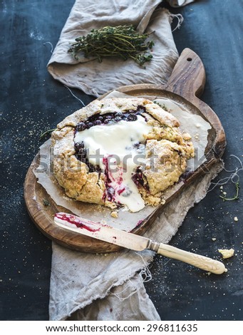 Homemade crusty pie or galette with blueberries, thyme and ice-cream on rustic wooden board over dark background