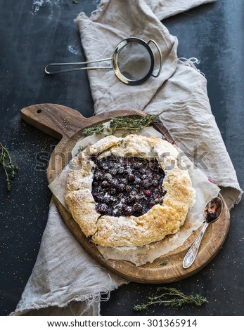 Homemade crusty pie or galette with blueberries, thyme and ice-cream on rustic wooden board over dark background