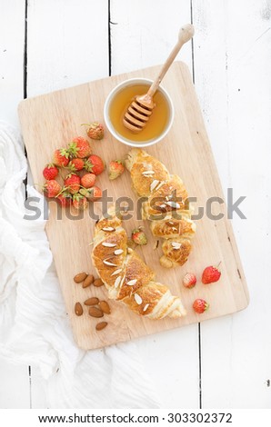 Freshly baked almond croissants with garden strawberries and honey on serving board over white rustic wood backdrop, top view