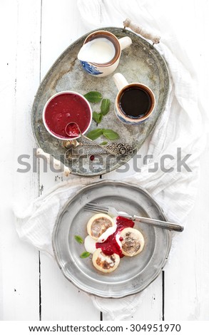 Rustic breakfast set. Russian cottage cheese pancakes or syrniki on a vintage metal plate with raspberry jam, sour cream and coffee. Rustic white backdrop, top view