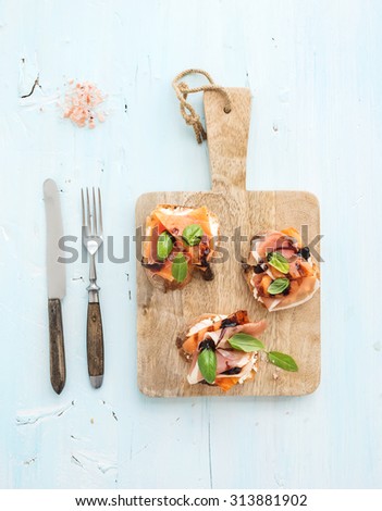 Bruschettas with Prosciutto, roasted melon, soft cheese and basil on wooden serving board over light blue background, top view