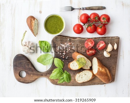 Pesto sauce, bread, cherry-tomatoes, fresh basil and garlic on rustic walnut chopping board over white wooden backdrop, top view