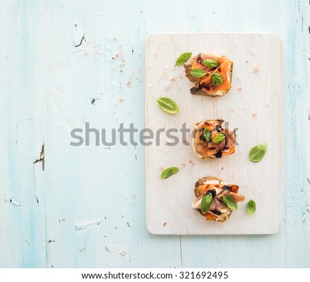 Bruschettas with Prosciutto, roasted melon, soft cheese and basil on white wooden serving board over light blue background, top view, copy space
