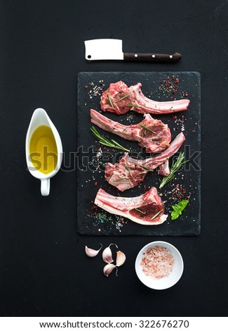 Raw lamb chops. Rack of Lamb with garlic, rosemary and spices on black slate tray, oil in a saucer, salt, dinnerware over dark rustic wood background