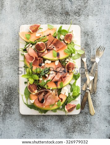 Prosciutto, melon, fig and soft cheese salad on a white serving board over grunge background, top view