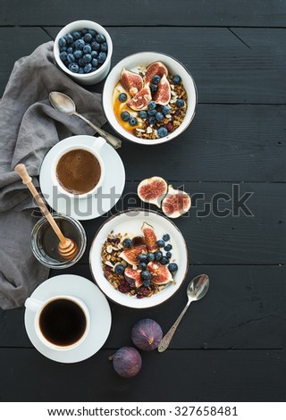 Healthy breakfast set. Bowls of oat granola with yogurt, fresh blueberries and figs, coffee, honey, over black wooden backdrop. Top view, copy space