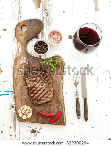 Grilled ribeye beef steak with herbs, spices  and glass of red wine on walnut cutting board over white rustic wooden background, top view