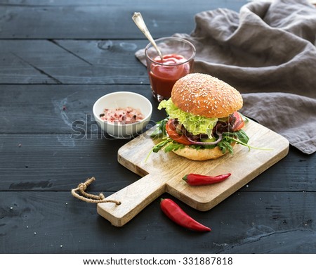 Fresh homemade burger on dark serving board with spicy tomato sauce, sea salt and herbs over dark wooden background, copy space