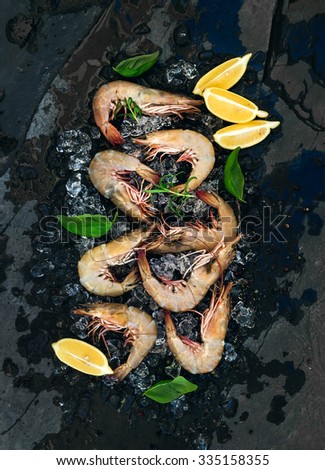 Fresh uncooked shrimps with lemon, herbs and spices on chipped ice over dark slate stone backdrop, top view