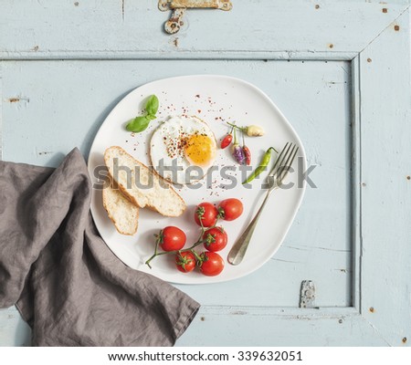 Breakfast set. Fried egg, bread slices, cherry tomatoes, hot peppers and herbs on white ceramic plate over light blue wooden backdrop, top view