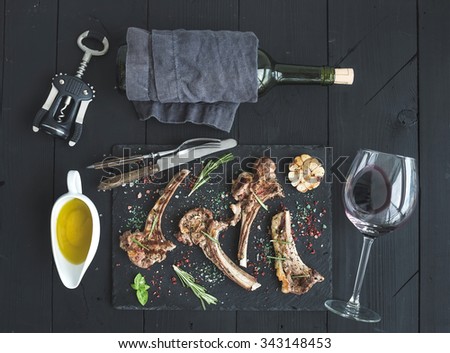 Grilled lamb chops. Rack of Lamb with garlic, rosemary and spices on slate tray, wine glass, oil in a saucer, corkscrew and bottle over black wood background. Top view
