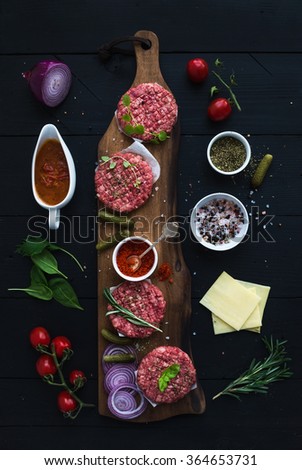 Ingredients for cooking burgers. Raw ground beef meat cutlets on wooden chopping board, onion, cherry tomatoes, greens, pickles, tomato sauce, cheese, herbs and spices over black background, top view