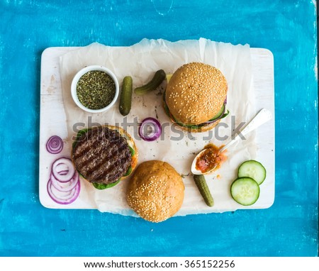 Fresh beef burger with cheese, vegetables and spicy tomato sauce on paper and white serving board, blue wooden background, top view