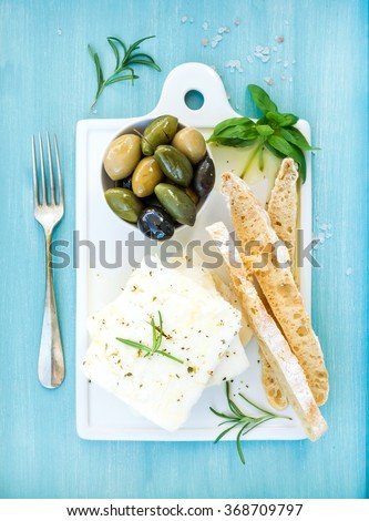 Fresh feta cheese with olives, basil, rosemary and bread slices on white ceramic serving board over bright turquoise blue painted wooden background, top view