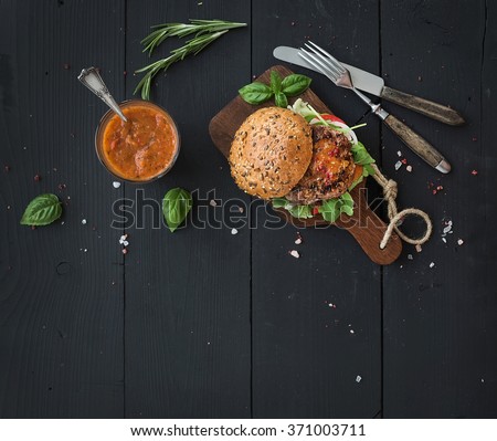 Fresh homemade burger on dark serving board with spicy tomato sauce, sea salt and herbs over dark wooden background. Top view, copy space, horizontal