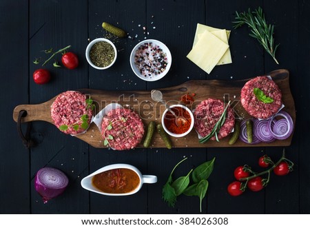 Ingredients for cooking burgers. Raw ground beef meat cutlets on wooden chopping board, red onion, cherry tomatoes, greens, pickles, tomato sauce, cheese, herbs and spices over black background