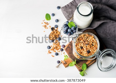 Healthy breakfast ingredients. Homemade granola in open glass jar, milk or yogurt bottle, blueberries and mint on white wooden background, top view, copy space